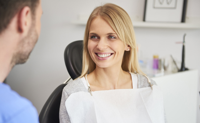 Female dentist in white coat smiling at male patient in modern dental office setting.