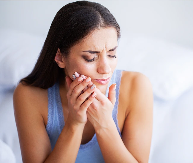 Woman experiencing tooth pain, holding her cheek with both hands against neutral background.