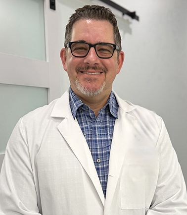 Professional male dentist wearing white lab coat and glasses, smiling in clinical office setting.